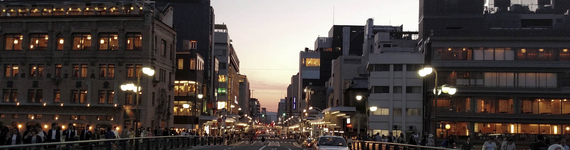 Calle de una ciudad al atardecer, iluminada, con tráfico y transitada